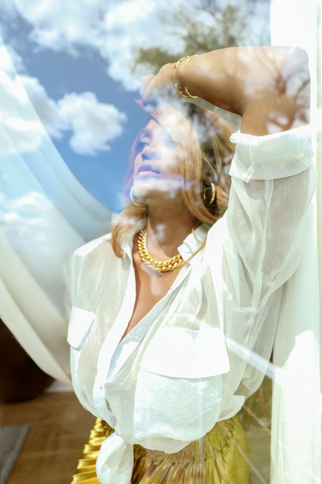 Toni Jones in a white blouse looking out through a window with clouds reflecting on the glass